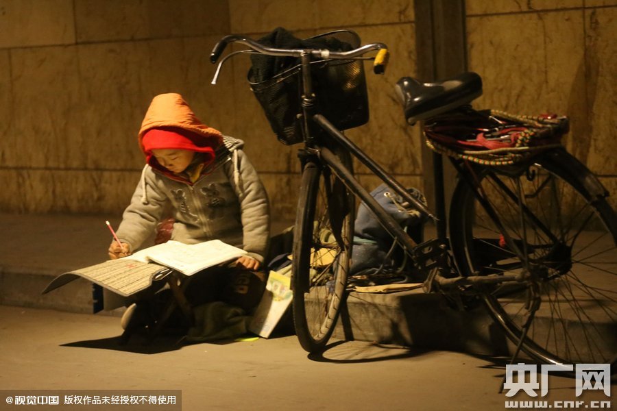 鄭州中原路京廣鐵路大橋下涵洞的馬路牙子邊，一位戴著紅色辣椒帽的小男孩趴在一個(gè)小馬扎上，上面鋪著一張車墊，在微弱的燈光下寫作業(yè)
