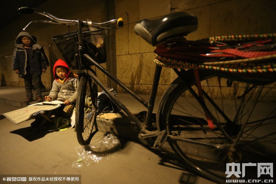 2015年11月8日晚，鄭州，7歲男孩涵洞橋下陪父母練攤，路邊燈下坐小馬扎上寫作業(yè)。