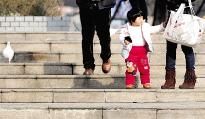 昨天中午，一女孩拉著家長的手在紫荊山公園曬太陽，氣溫升高，孩子的棉衣解開了扣子。